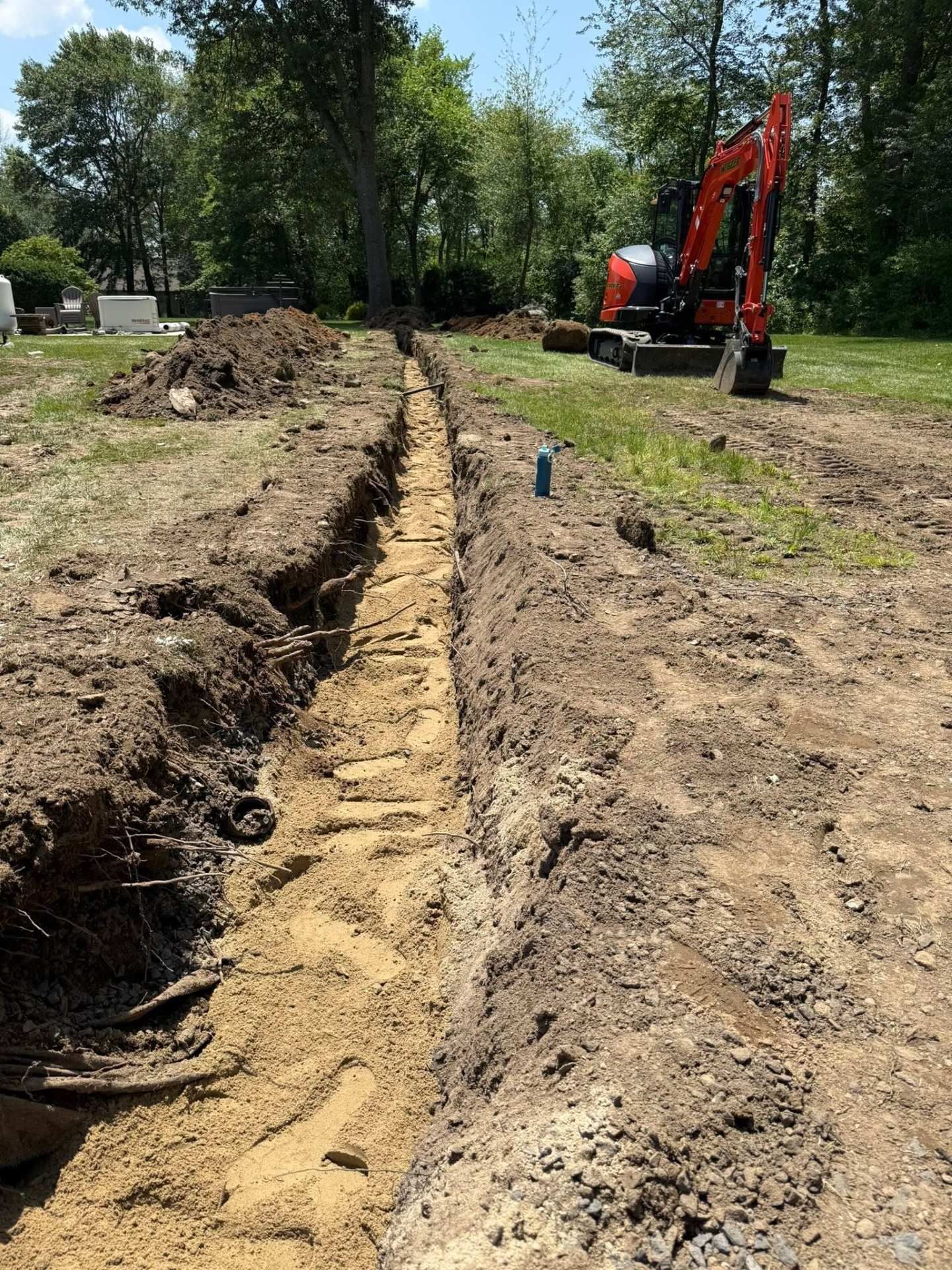 Excavator digging a long trench in a dirt field beside a grassy area and trees