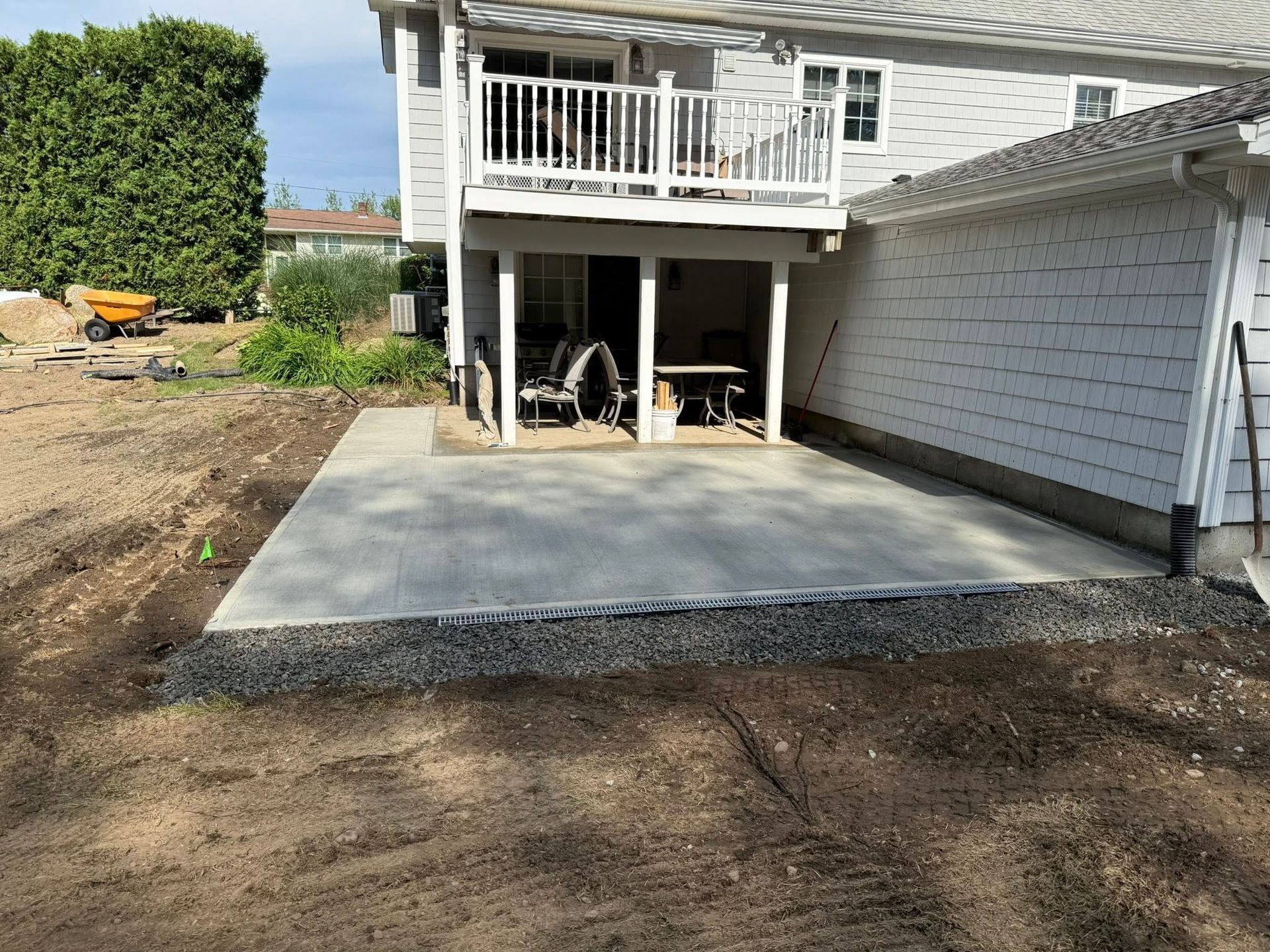Backyard concrete patio under a raised deck beside a house, with dirt yard and hedge nearby