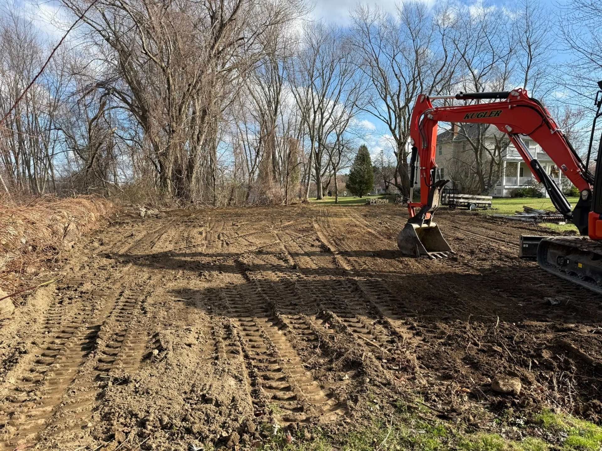 Red excavator grading a muddy clearing beside leafless trees under a blue sky.