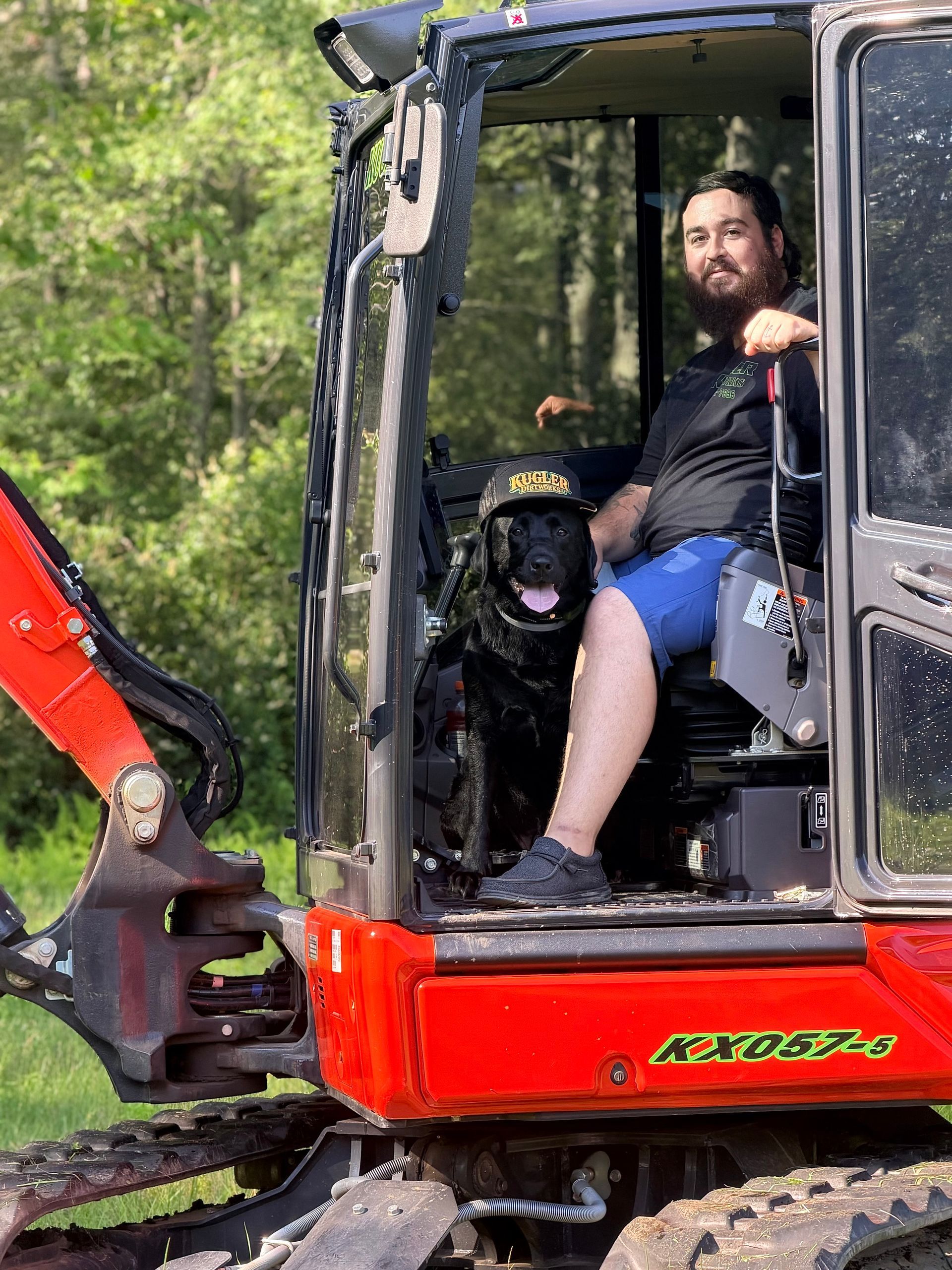 Man seated in a red excavator cab with a black dog beside him, outdoors among trees