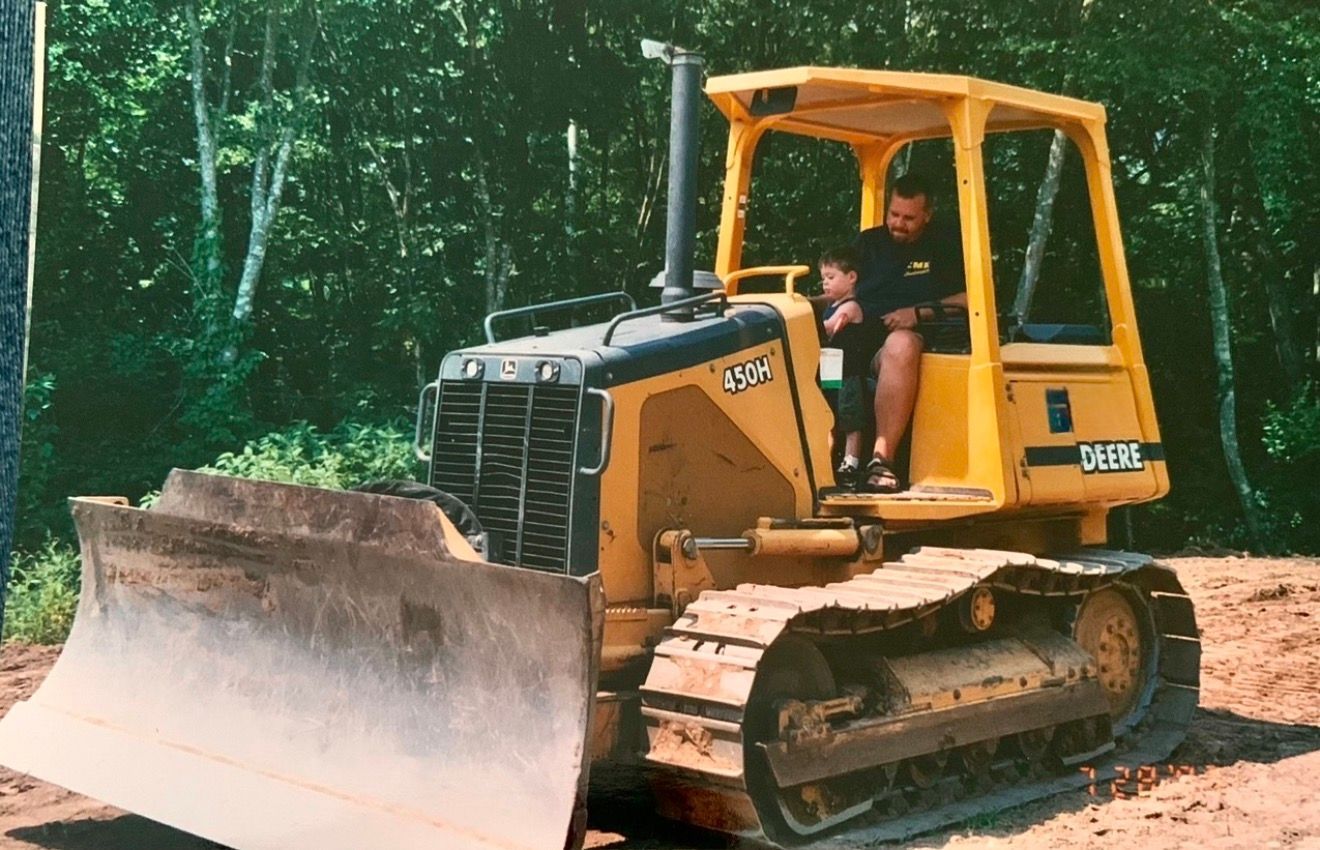 Yellow bulldozer on a dirt road in a forest, with a driver visible in the cab.