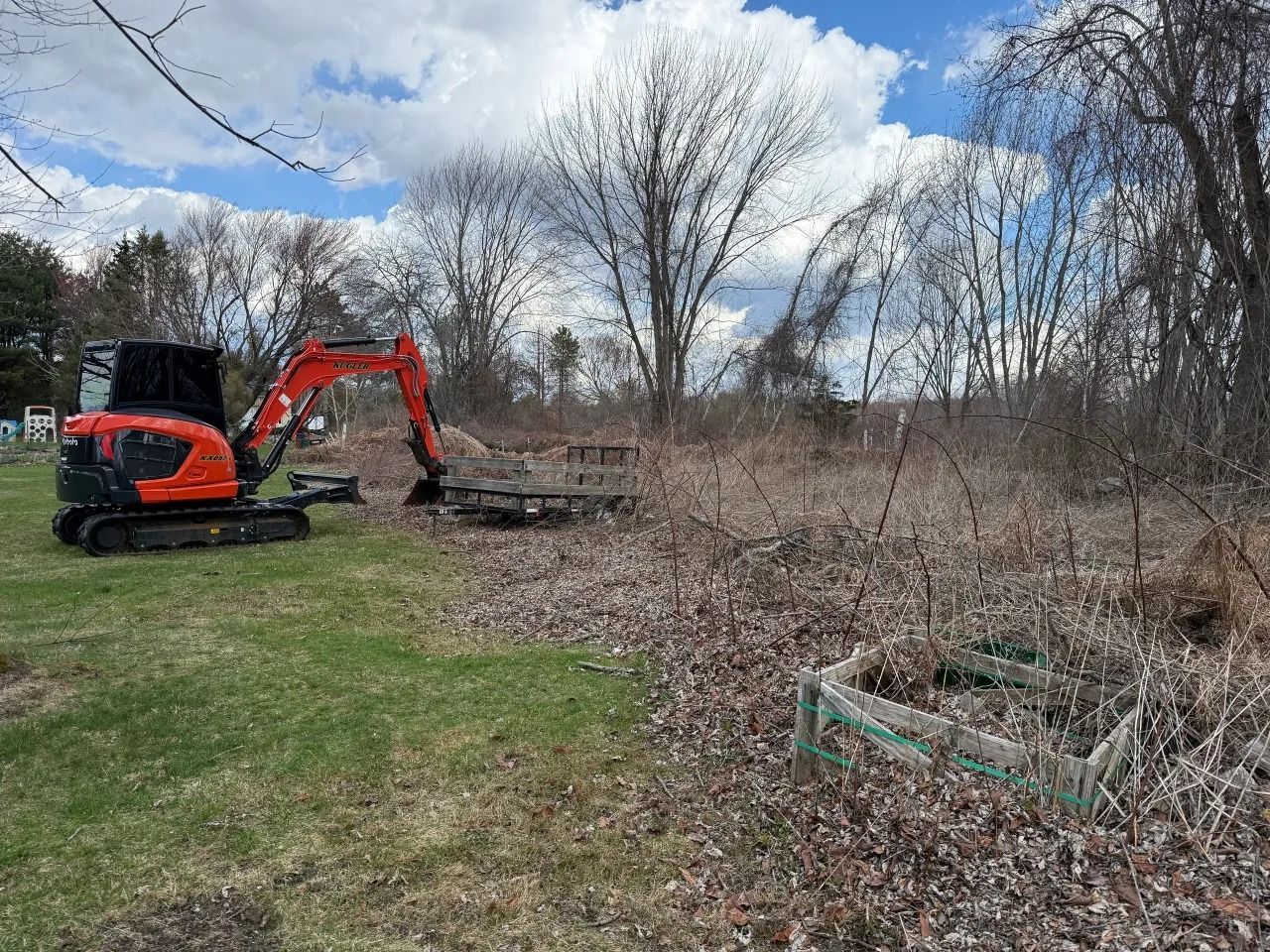 Orange excavator clearing brush in a grassy field under a cloudy sky