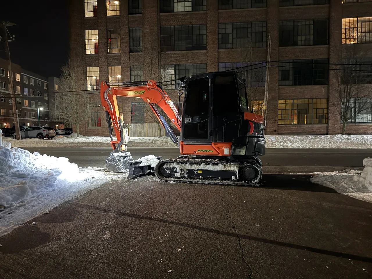 Orange excavator clearing snow on a wet street at night beside an apartment building