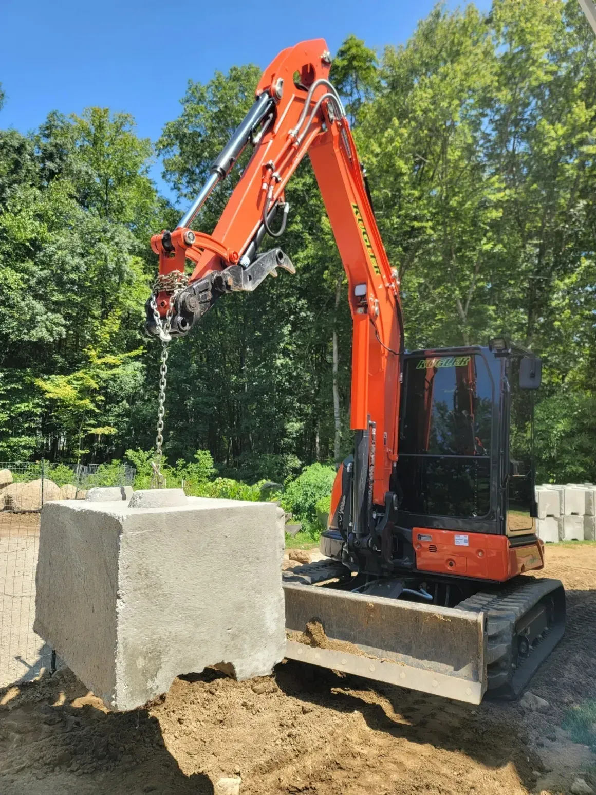 Orange excavator lifting a concrete block at a construction site surrounded by trees