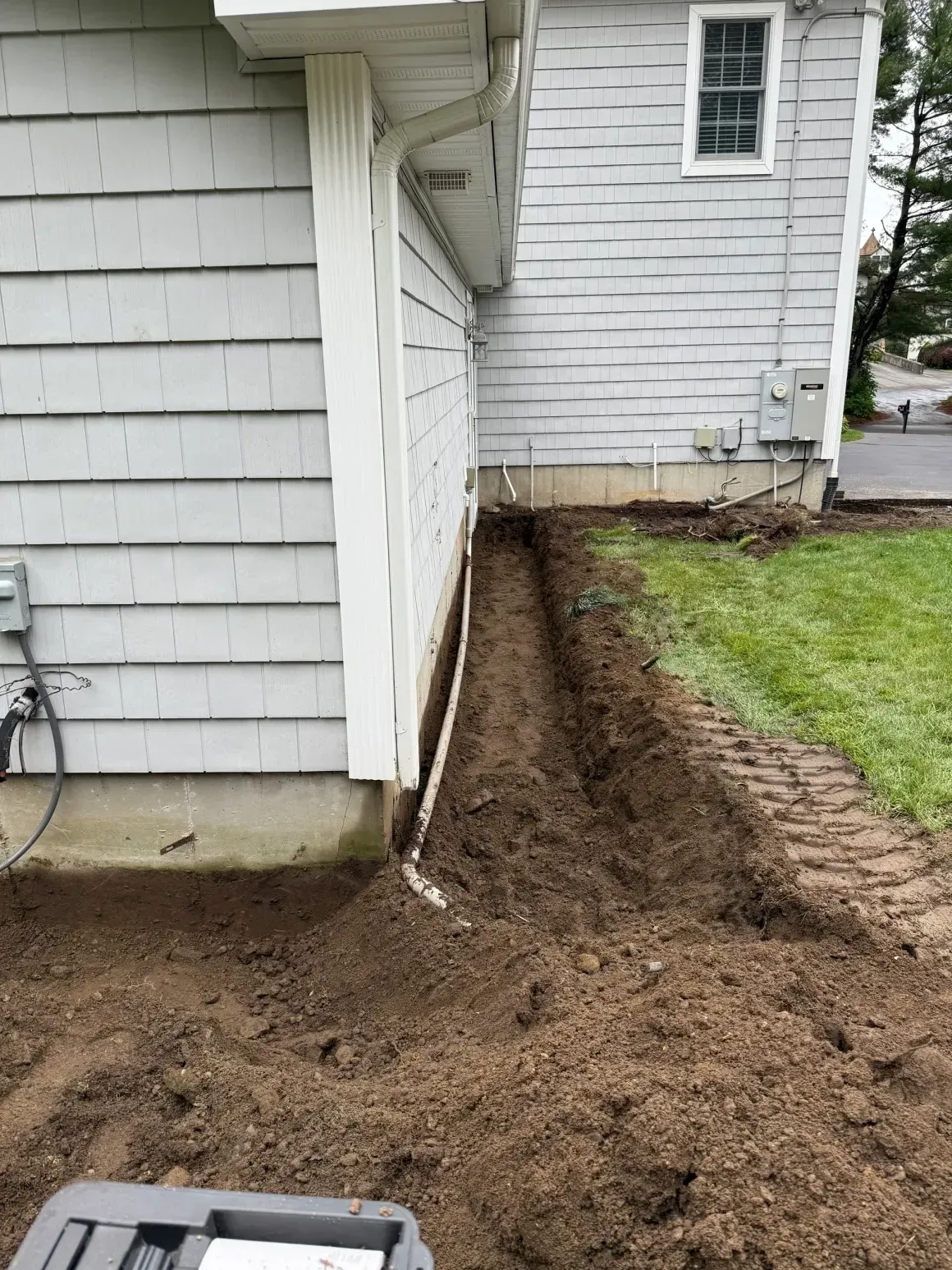 House side with fresh dirt trench beside the foundation and a patch of lawn.