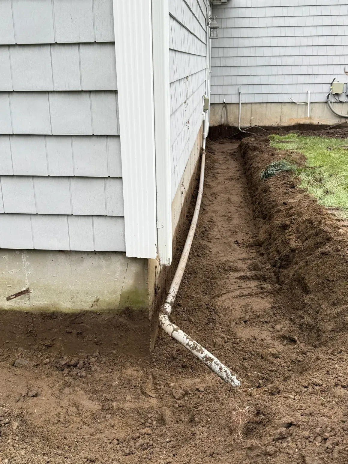 Foundation repair trench along house siding with exposed drain pipe and fresh dirt.
