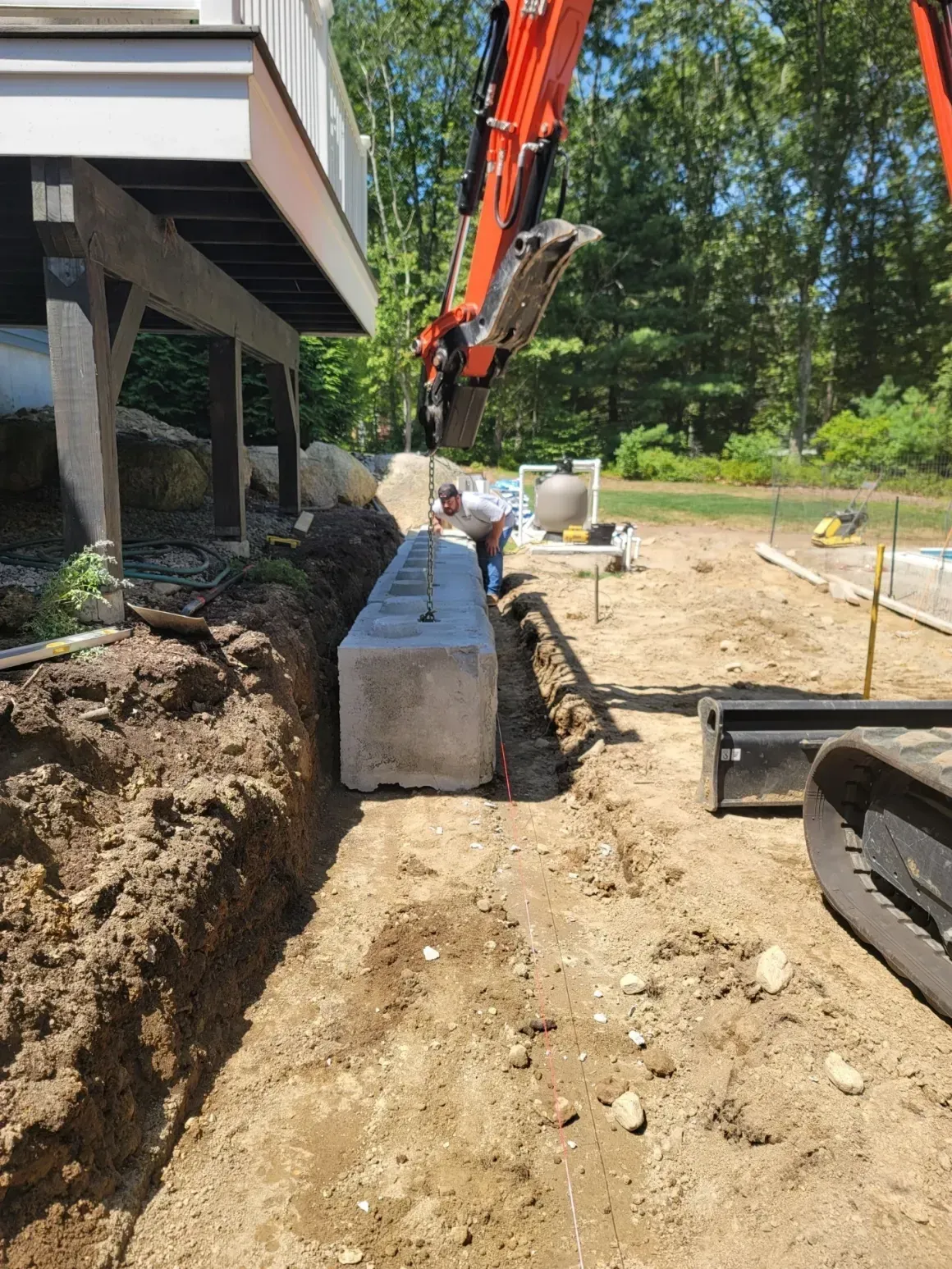 Construction site with excavator placing concrete blocks beside a house foundation and dirt trench