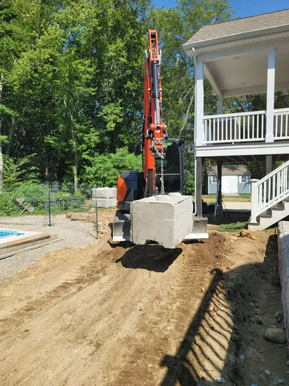 Orange drilling rig hoists a concrete block beside a white house on a dirt construction site.