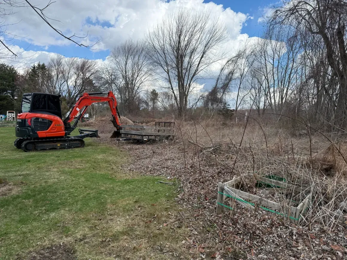 Orange excavator clearing brush in a grassy field under a cloudy sky