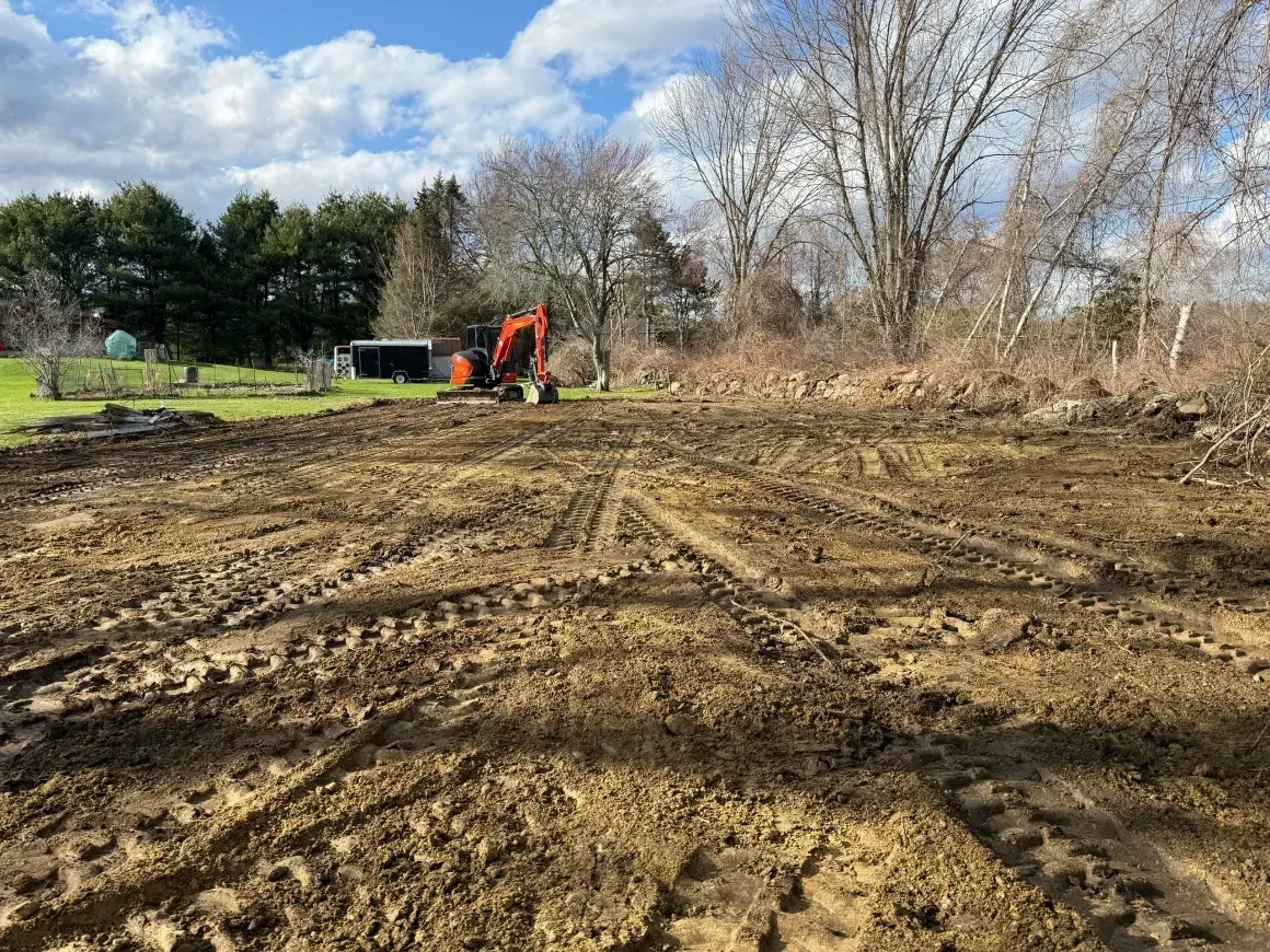 Cleared muddy lot with excavator working under a cloudy sky and trees in the background