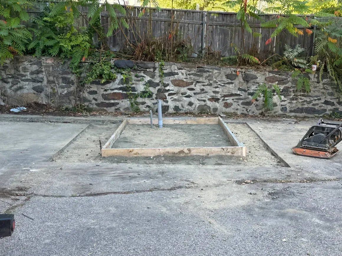 Small concrete pad with a square wooden frame and vertical pipe, beside a stone wall and garden fence.