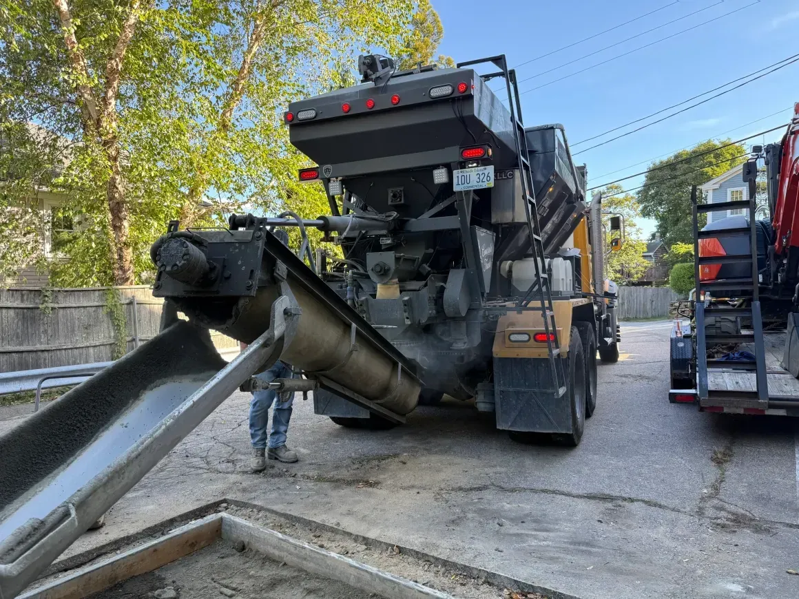 Road paving machine spreading asphalt on a street, with workers nearby and a dump truck at right.