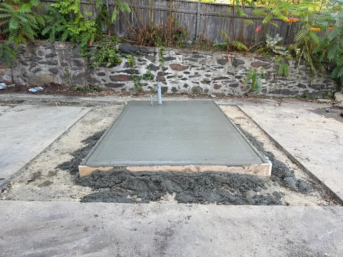 Freshly poured concrete slab framed by rough gravel, with a stone wall and fence in the background