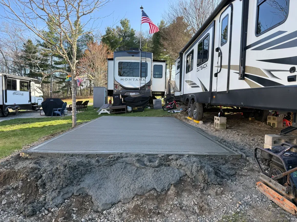 Damaged concrete RV pad between parked trailers in a campground, with a cracked edge in front.