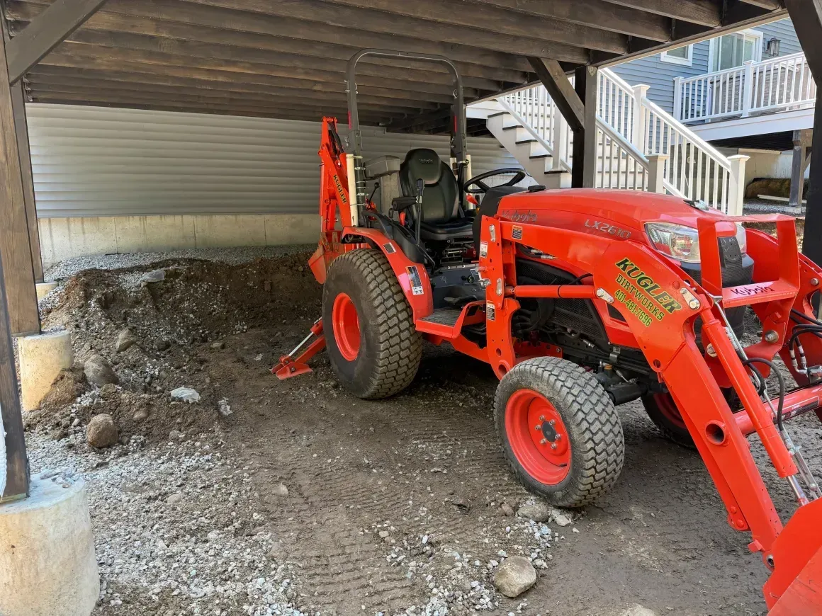 Red tractor with front loader under a deck beside a dirt pile and stairs