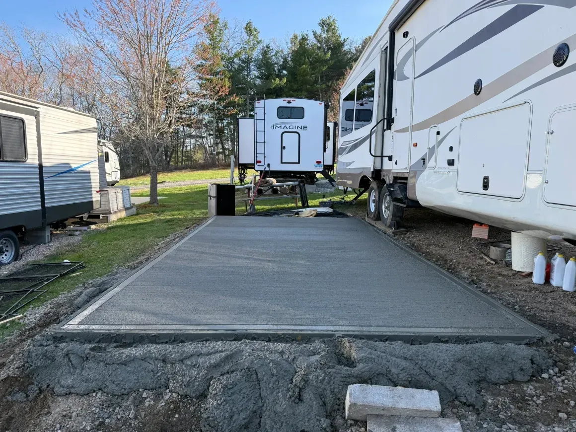 RVs parked beside a newly poured concrete pad in a grassy campground area.