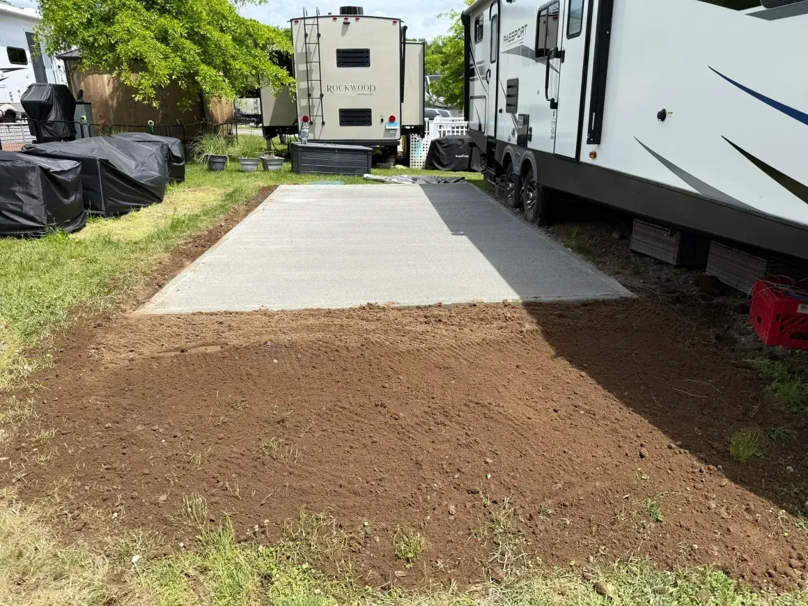 RV park pad with fresh dirt, a concrete driveway, and large parked trailers on both sides