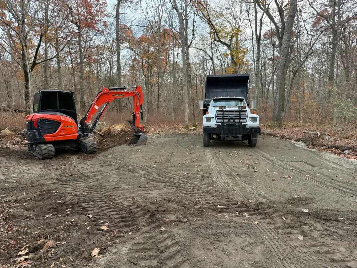 Orange excavator and white dump truck on a dirt path in a leafless wooded area