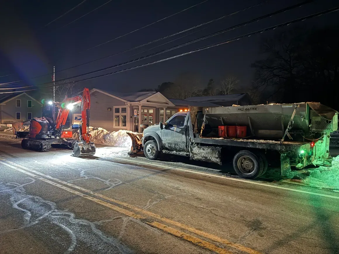 Snowy nighttime roadwork with a red excavator loading a white dump truck on a residential street