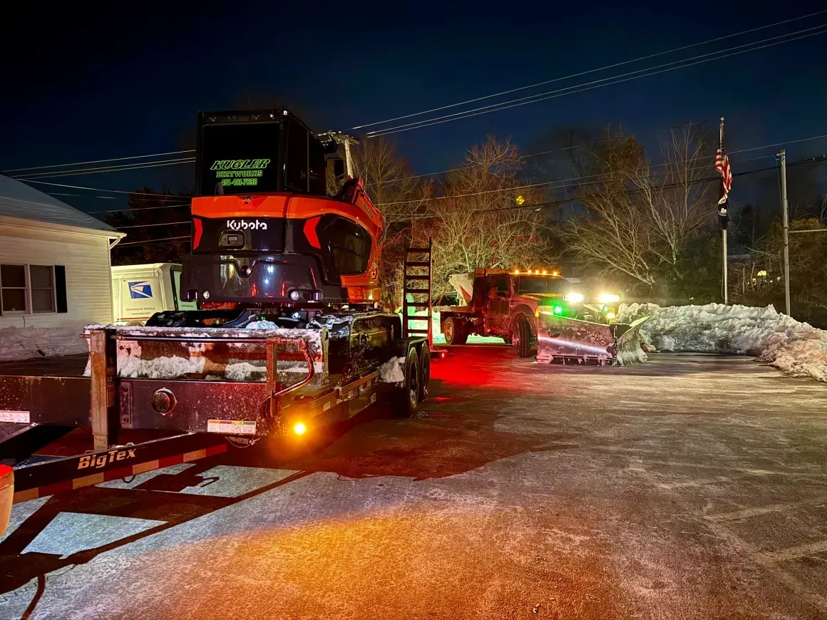 Red tow truck with flashing lights on a snowy street at night, near a house and an American flag.