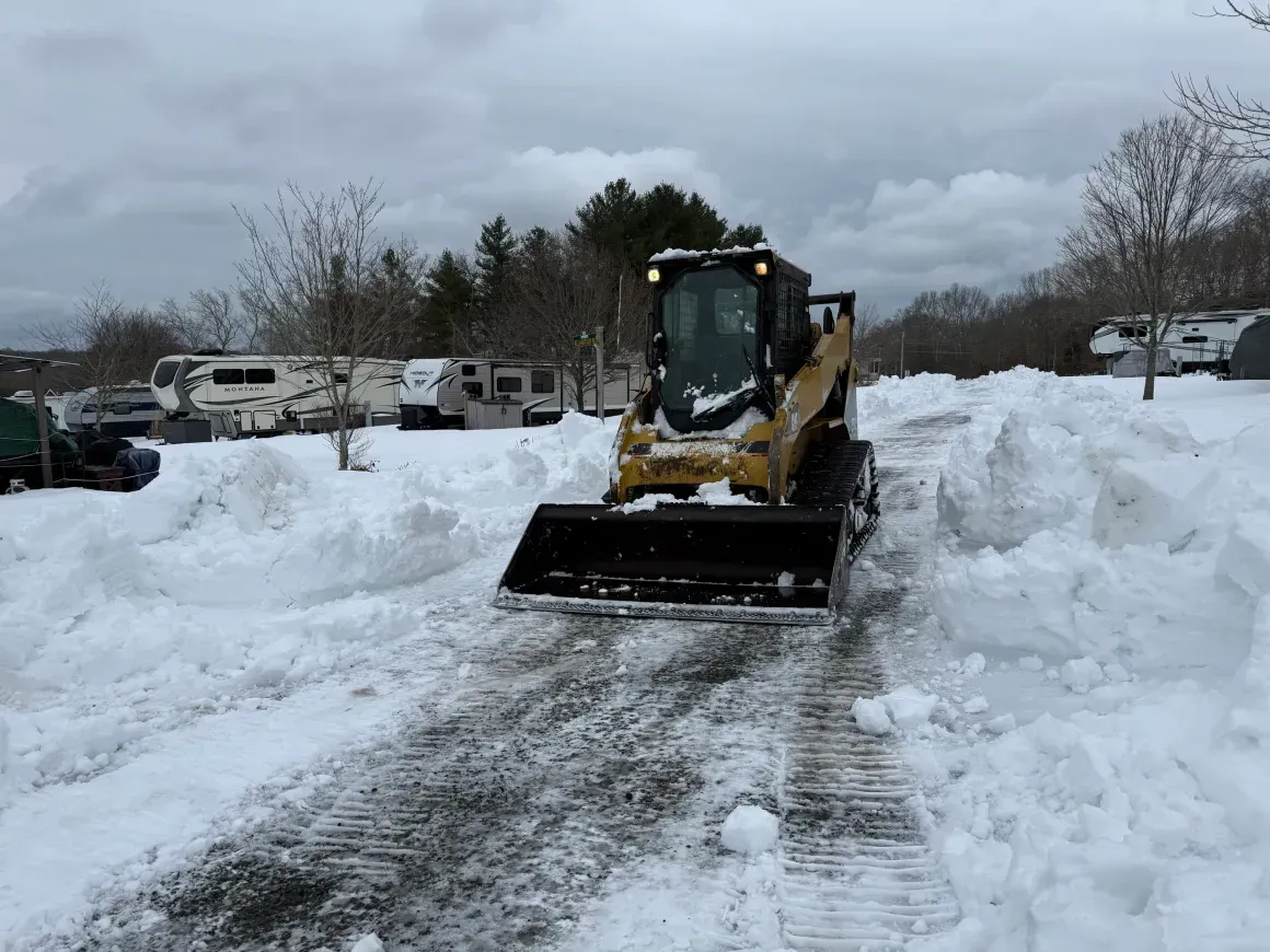 Yellow snowplow clearing a snow-covered driveway under a cloudy sky
