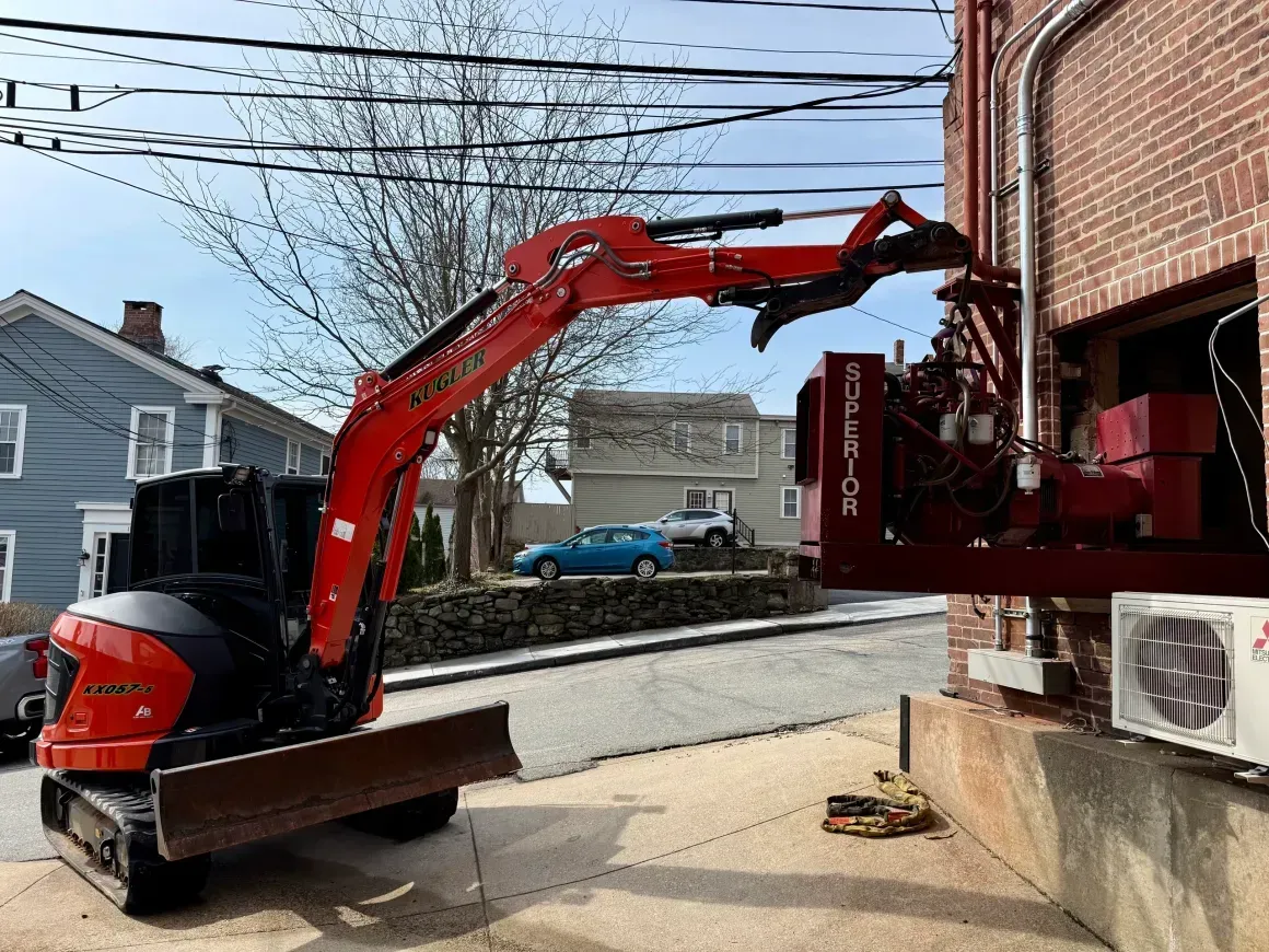 Red excavator lifting debris into a dumpster beside a brick building on a street