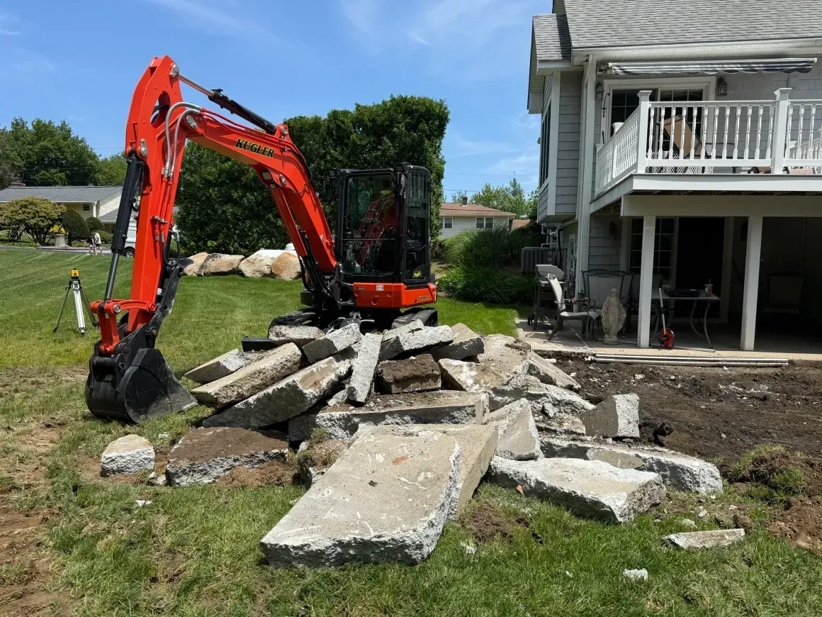 Orange excavator demolishing broken concrete slabs beside a house yard