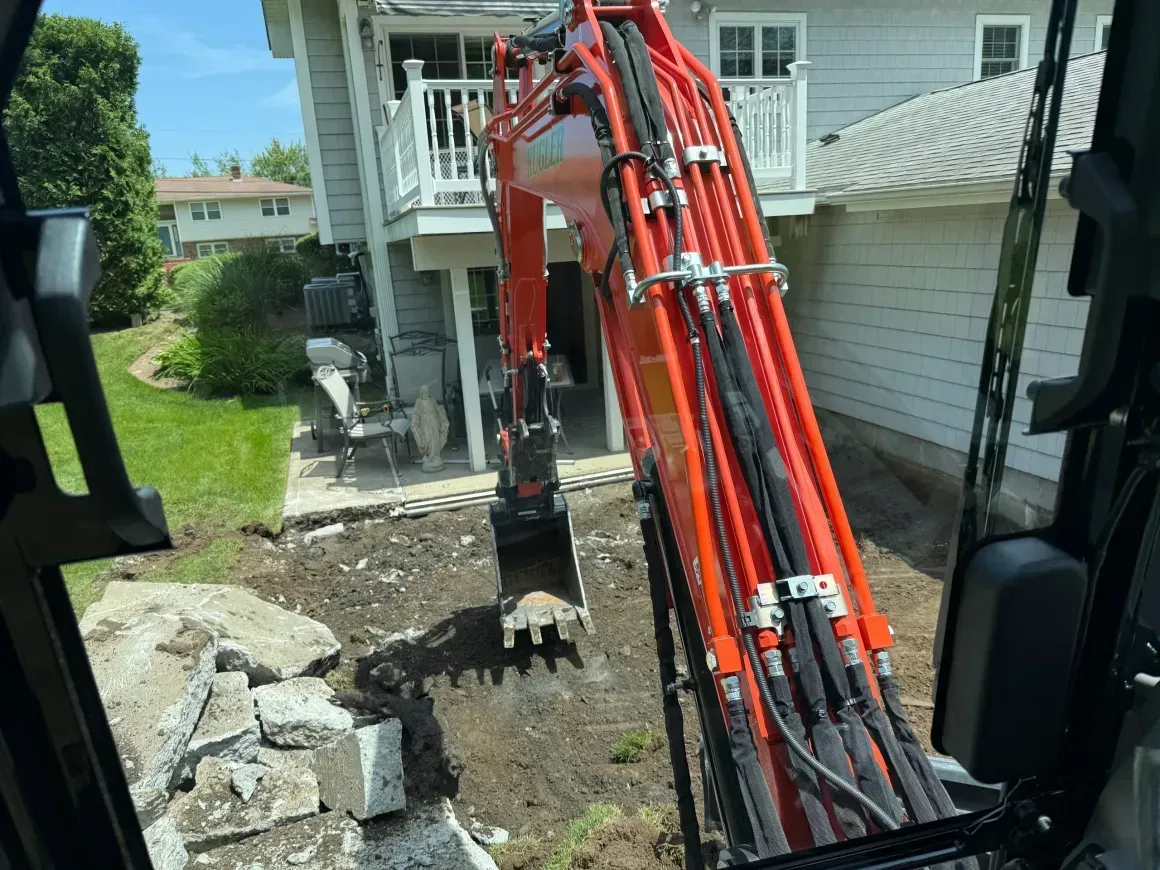 Excavator digging a trench beside a house, with red hydraulic arm and piled dirt and rocks.