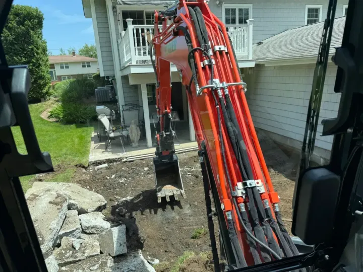 Excavator digging a trench beside a house, with red hydraulic arm and piled dirt and rocks.