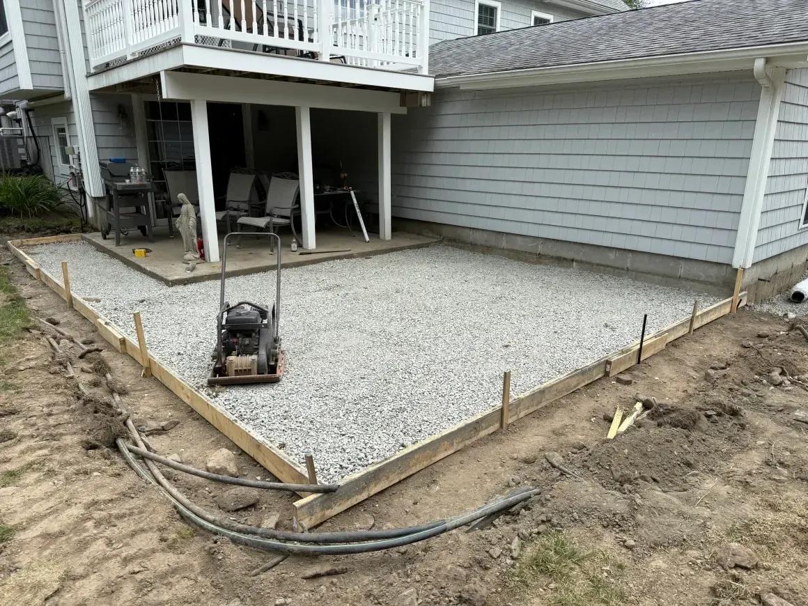 Gravel patio area under a deck beside a gray house, with edging stakes and a compactor on the ground.