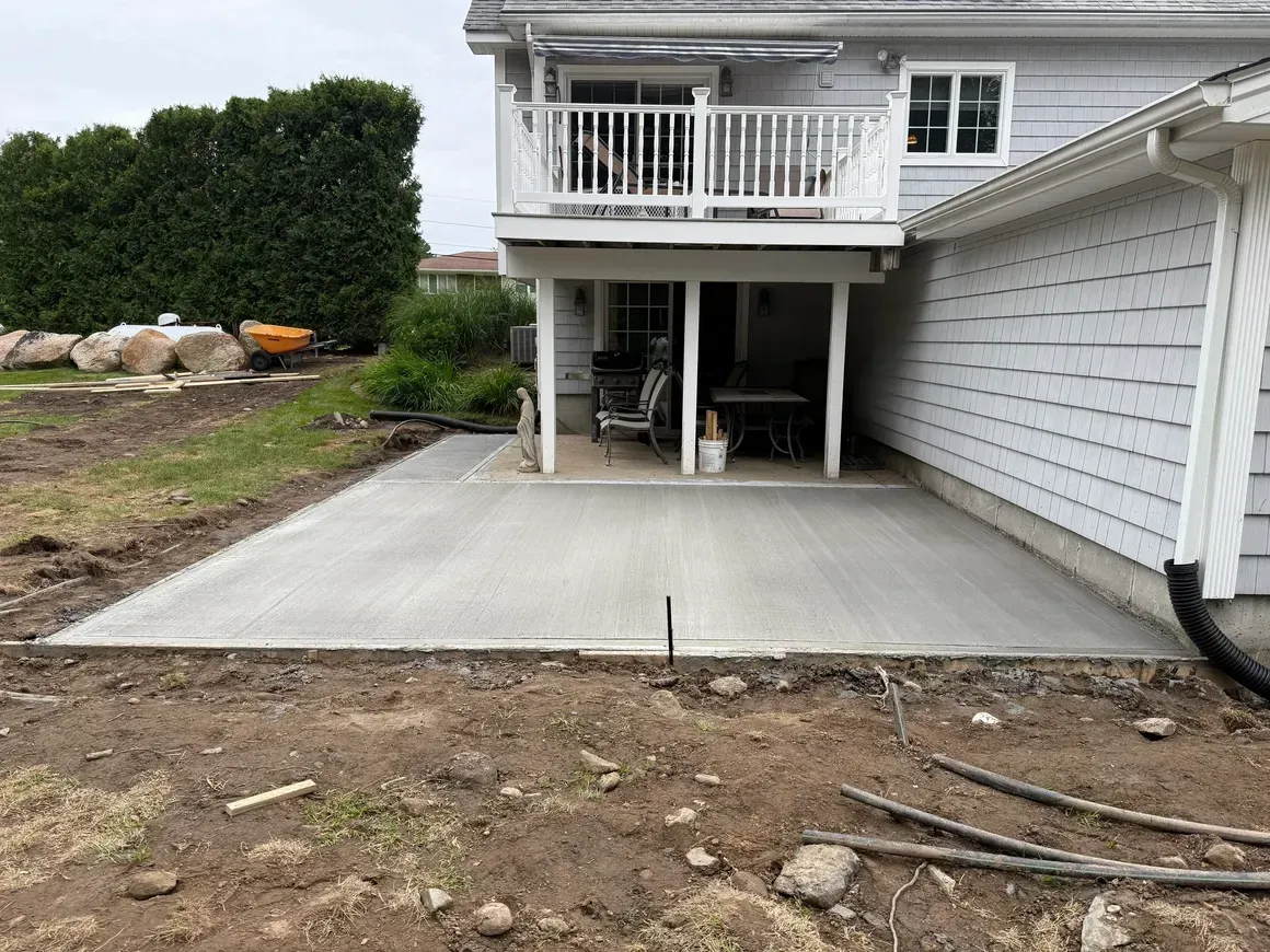 Concrete backyard patio beside a house with an upper deck and stairs, next to a white garage