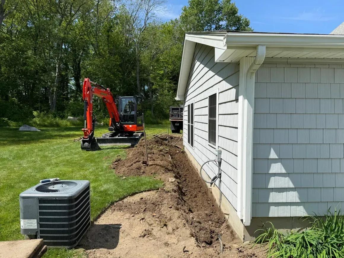 Mini excavator digging a trench beside a white house, with an HVAC unit in the foreground.