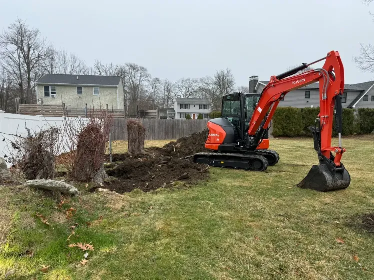 Orange excavator digging a trench in a grassy yard beside a fence and houses
