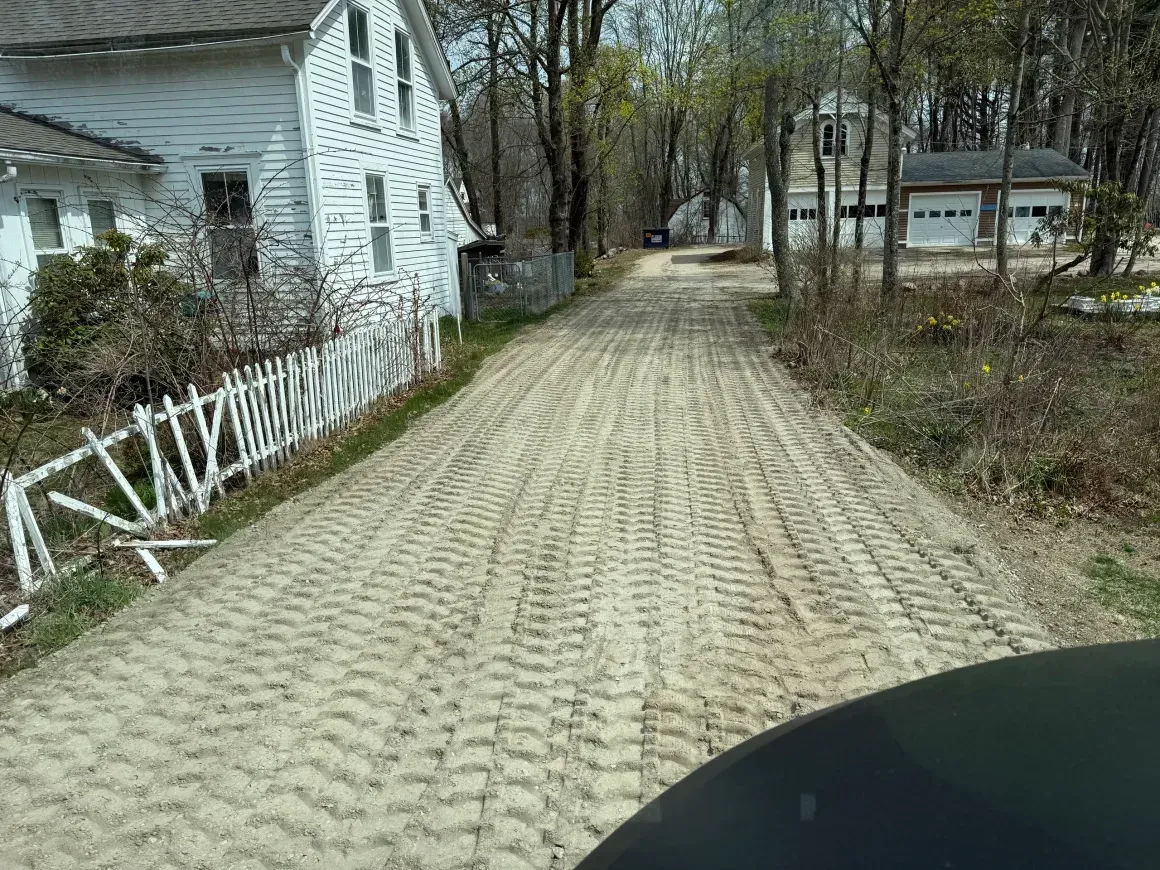 Paved driveway beside a white house, leading through a wooded yard on a cloudy day