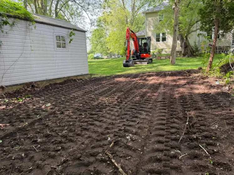 Red excavator grading a dirt yard beside a white shed in a wooded backyard.