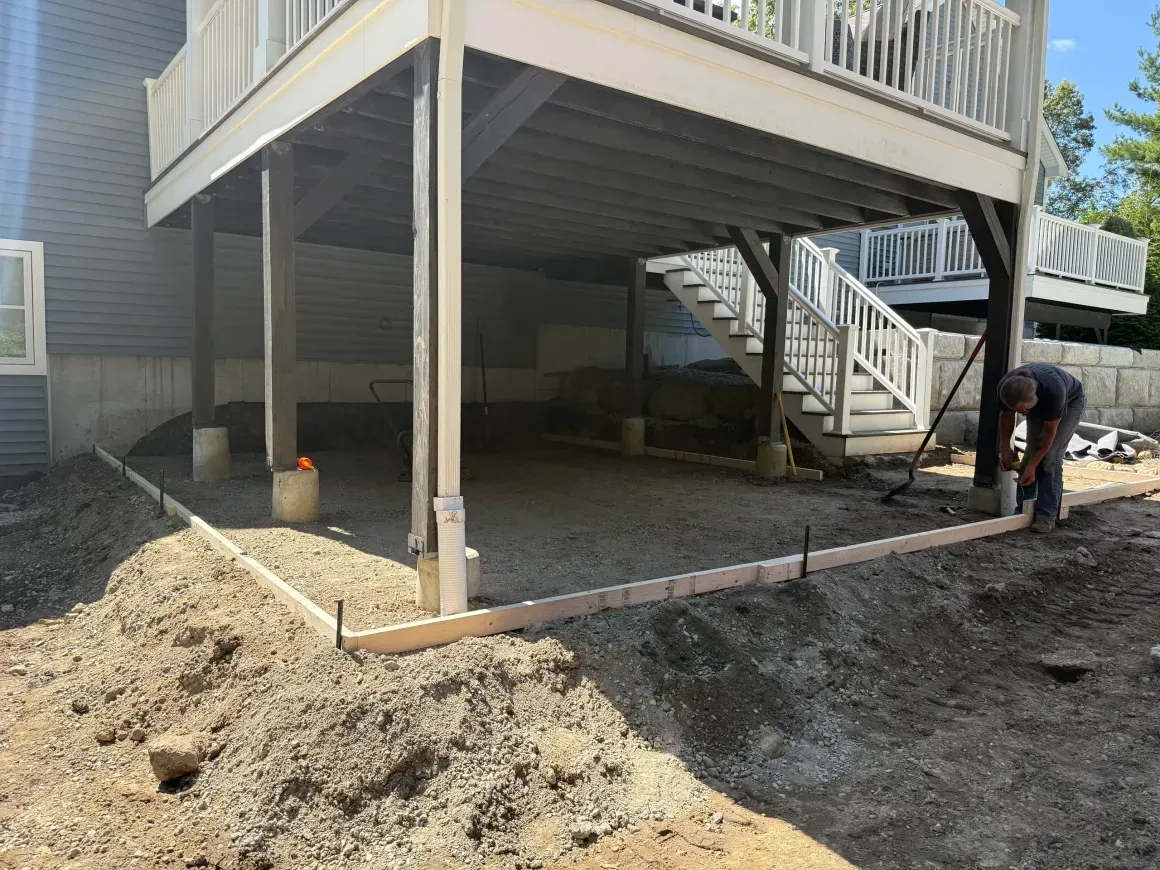 Raised deck under construction with exposed posts and stairs, and a worker leveling soil around the base.