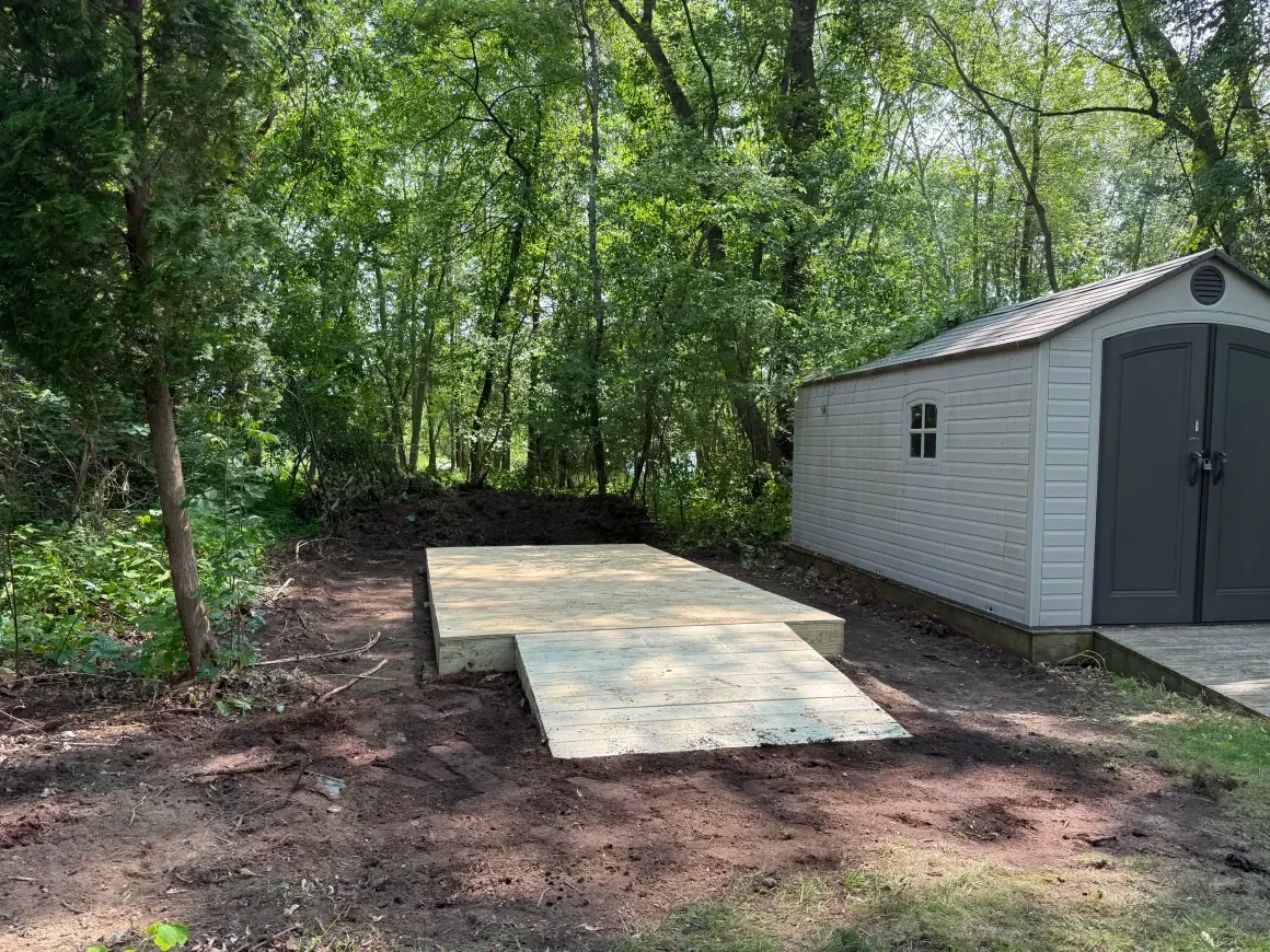 Wooden ramp beside a white shed in a wooded yard, with dirt ground and trees in the background