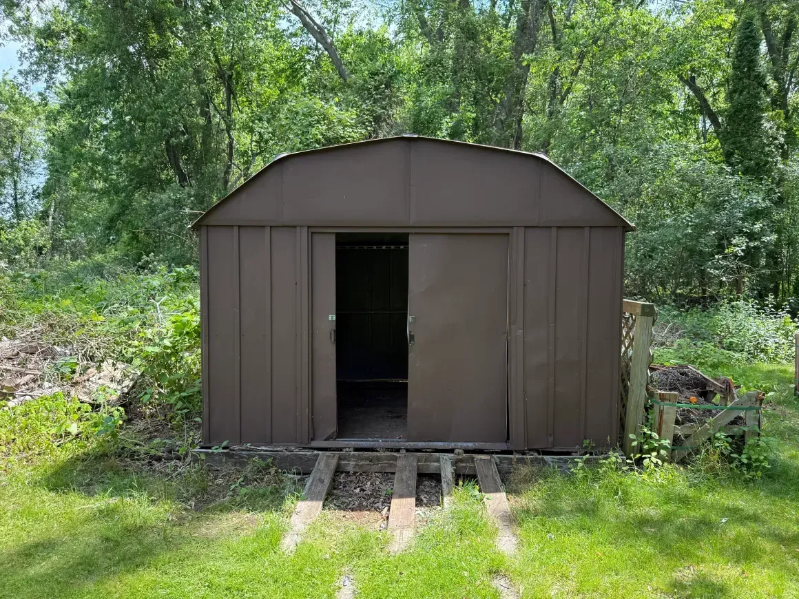 Brown metal shed with an open doorway in a wooded backyard, with a short ramp leading up to it