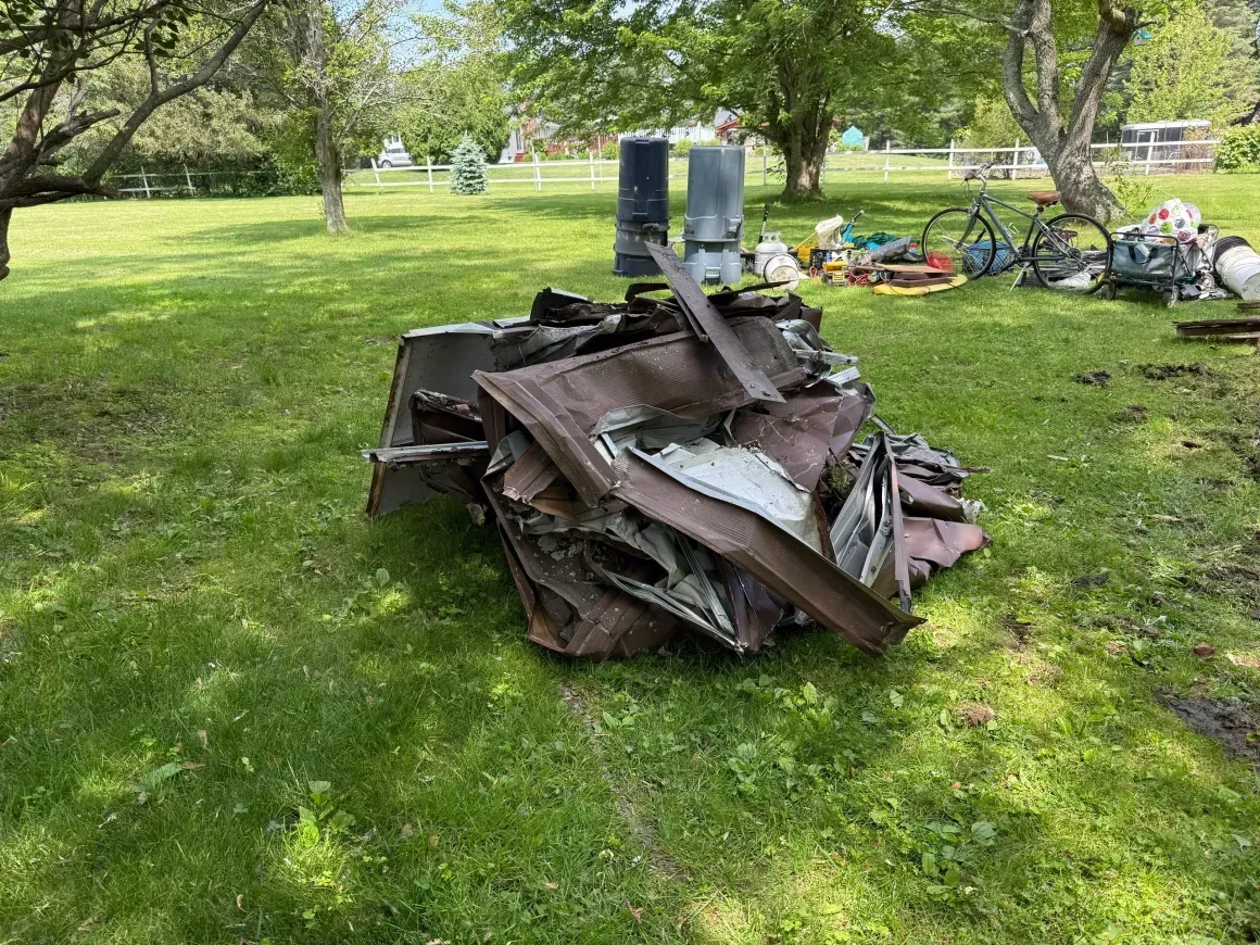 Pile of rusted scrap metal on grass in a sunny park with trees and scattered debris nearby