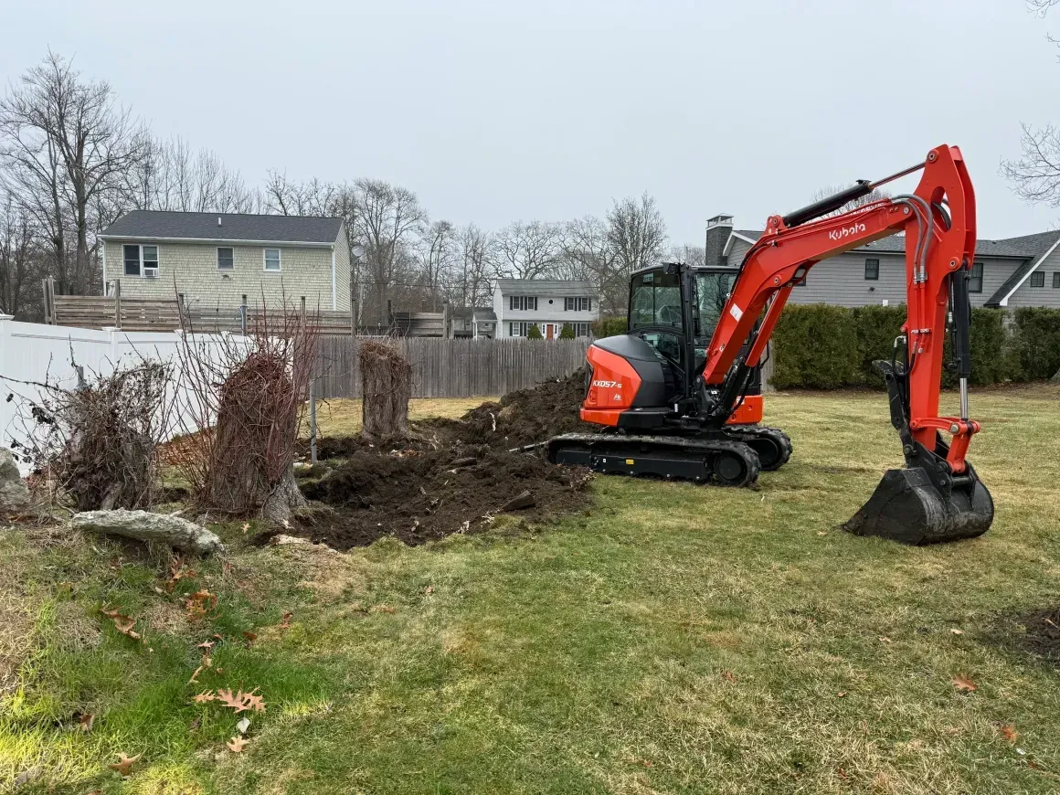 Orange mini excavator clearing soil in a backyard beside a fence and house