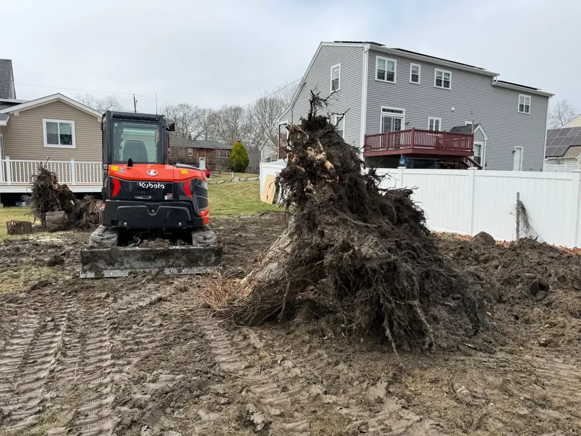 Red utility vehicle in muddy yard beside large pile of uprooted brush and a house under overcast sky