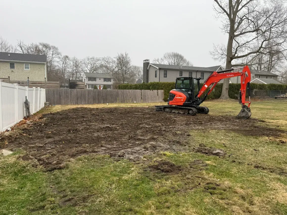 Orange excavator leveling a muddy backyard beside a fence and trees
