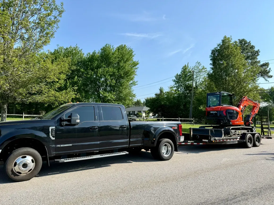 Black pickup towing a trailer with a red tractor on a tree-lined roadside