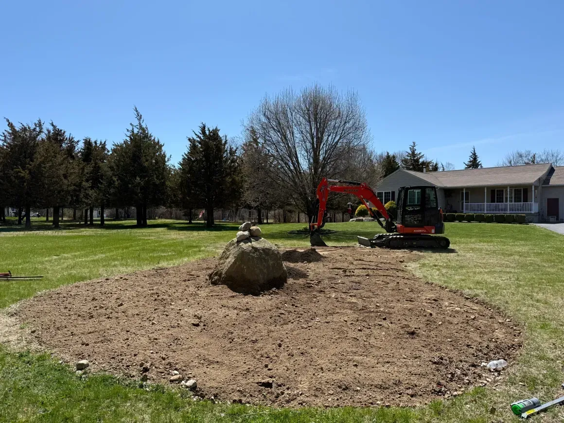 Excavator leveling soil around a mound in a grassy yard near a house and trees under a blue sky
