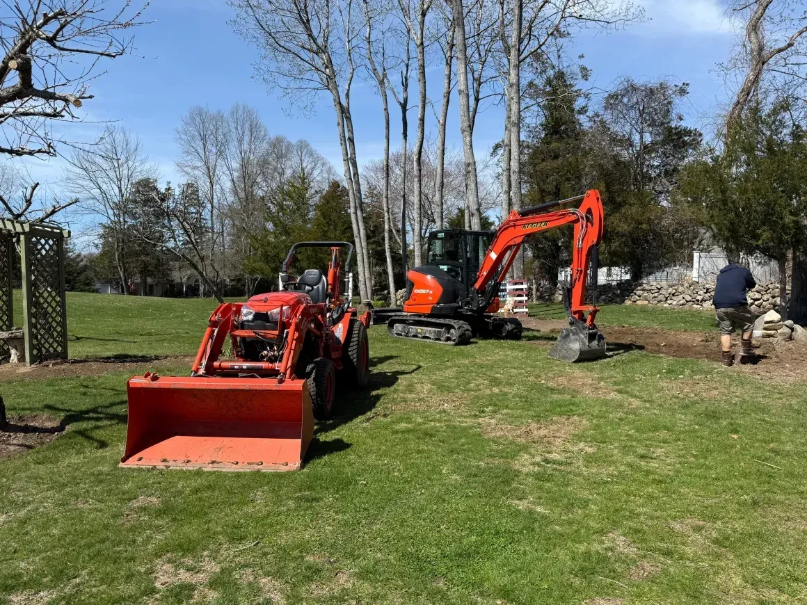 Red tractor and orange excavator on a grassy yard with a person nearby and trees in the background