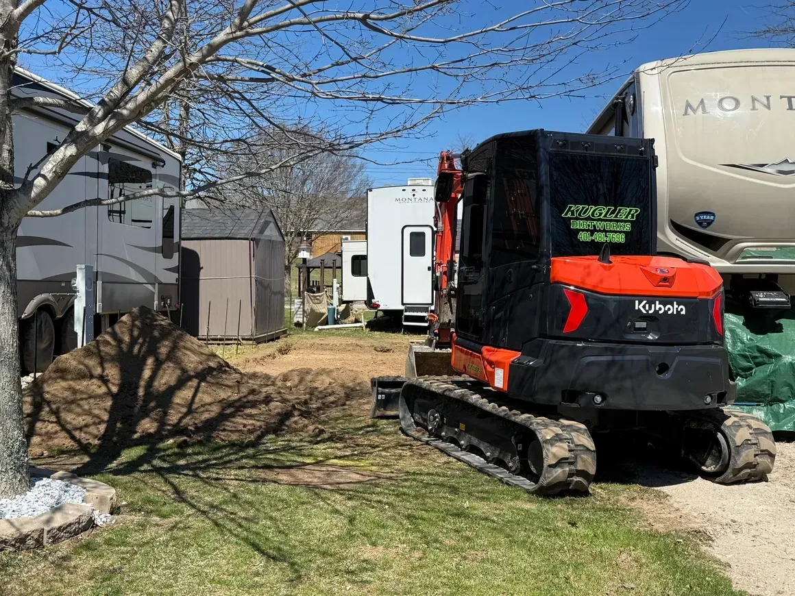 Black Kubota mini excavator parked beside RVs on a gravel lot under a blue sky