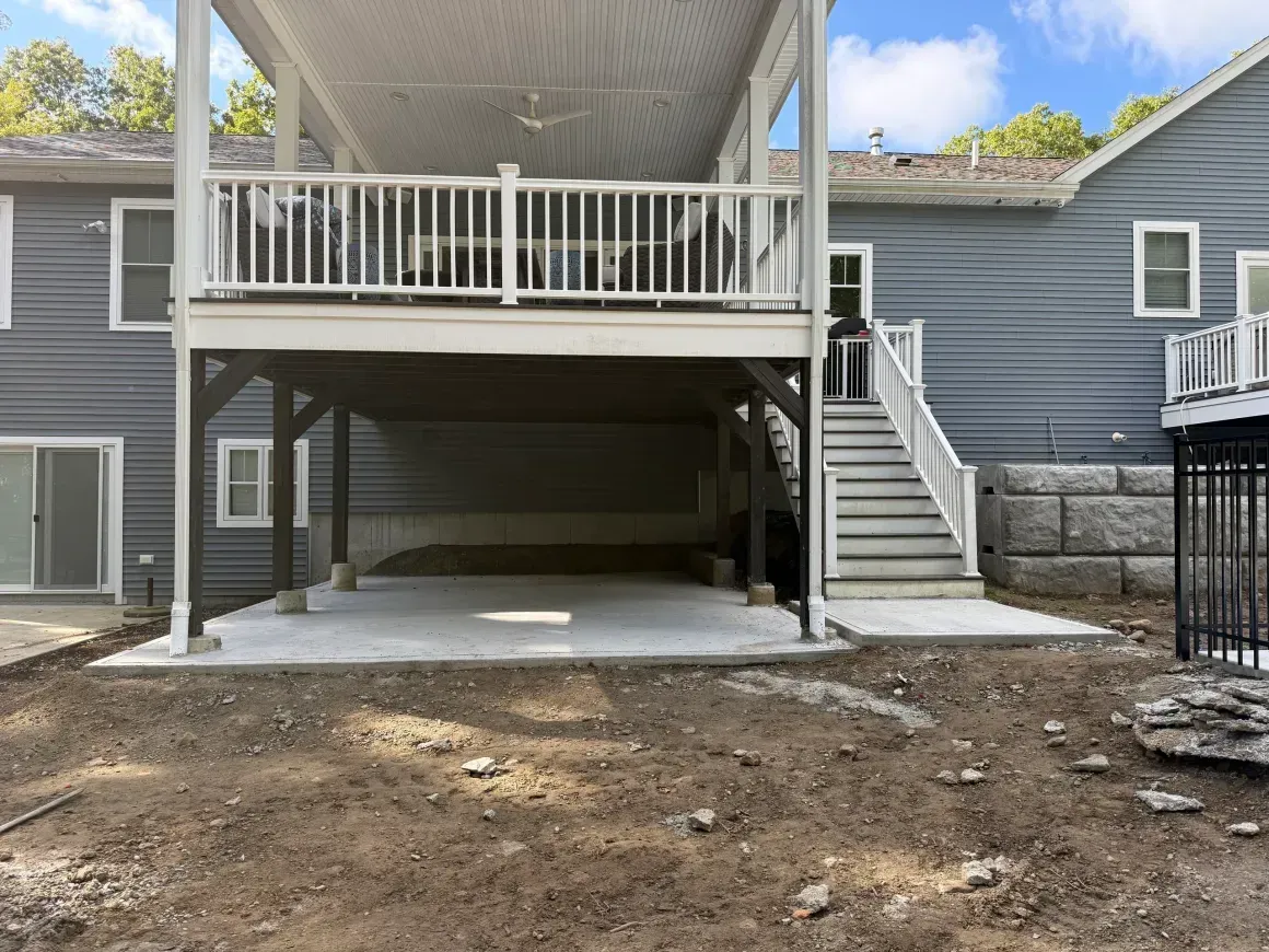 Covered patio and under-deck parking area behind a gray house, with stairs on the right and dirt foreground.