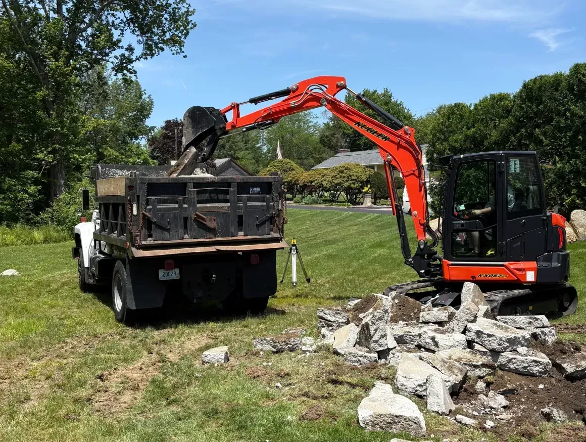 Mini excavator loading rubble into a dump truck in a grassy field under blue sky