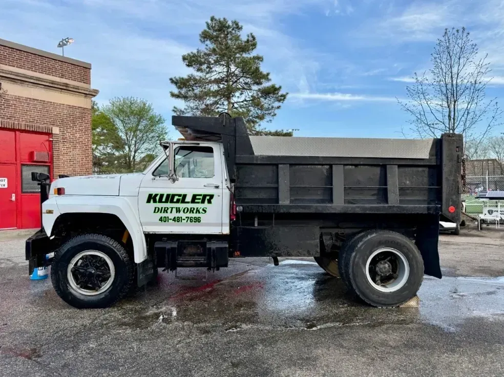 White dump truck with “Hugler Enterprises” logo parked beside a brick building on a wet lot