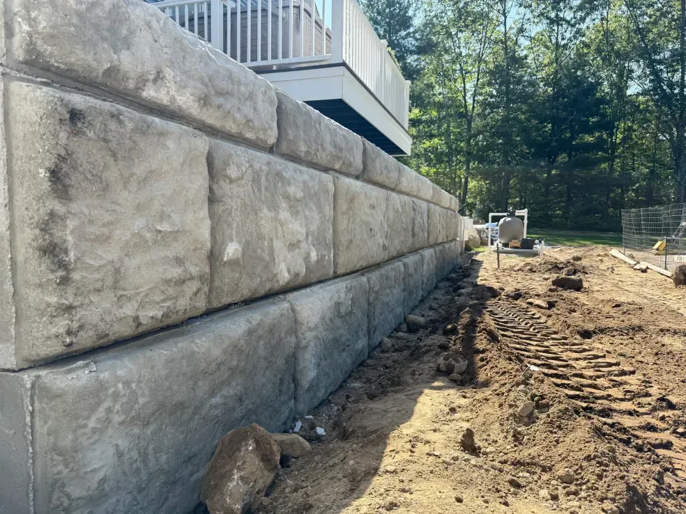 Stone retaining wall beside a dirt excavation site with a deck and trees in the background